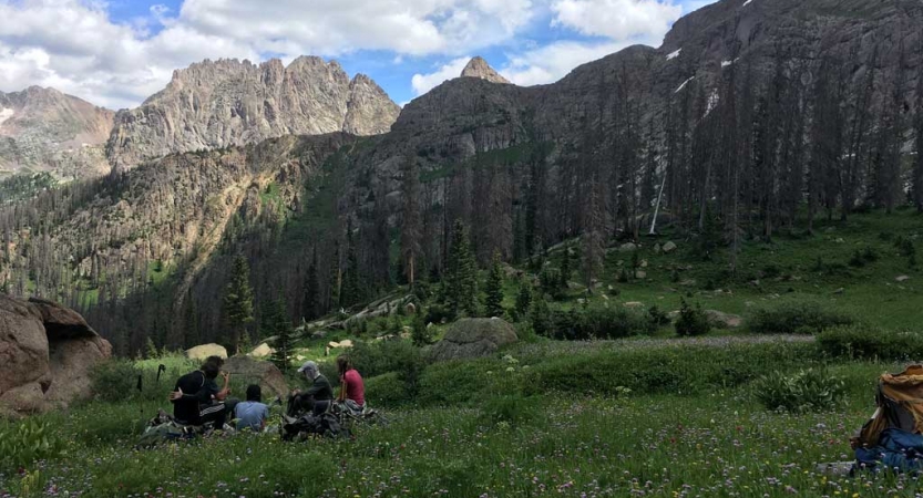 A group of people sit in green grass framed by rocky mountains. 